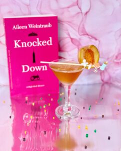 Knocked Down book with its Booktail in a martini glass containing amber liquid with a baby themed stirrer.